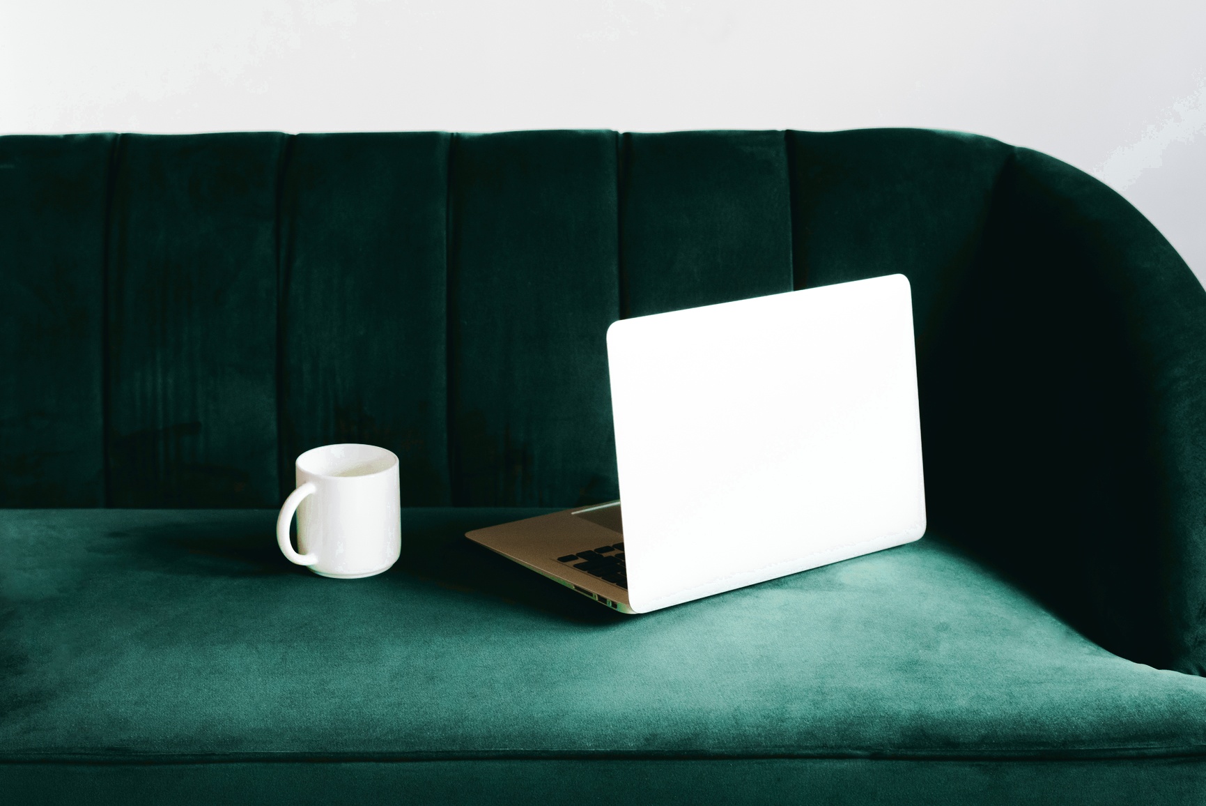 A laptop and coffee cup on a teal velvet banquette — the work, without the worker.